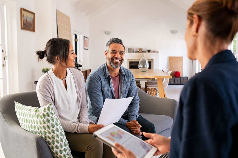 A woman sits with a couple in their home and is explaining something to them. She has an iPad and they are holding some papers