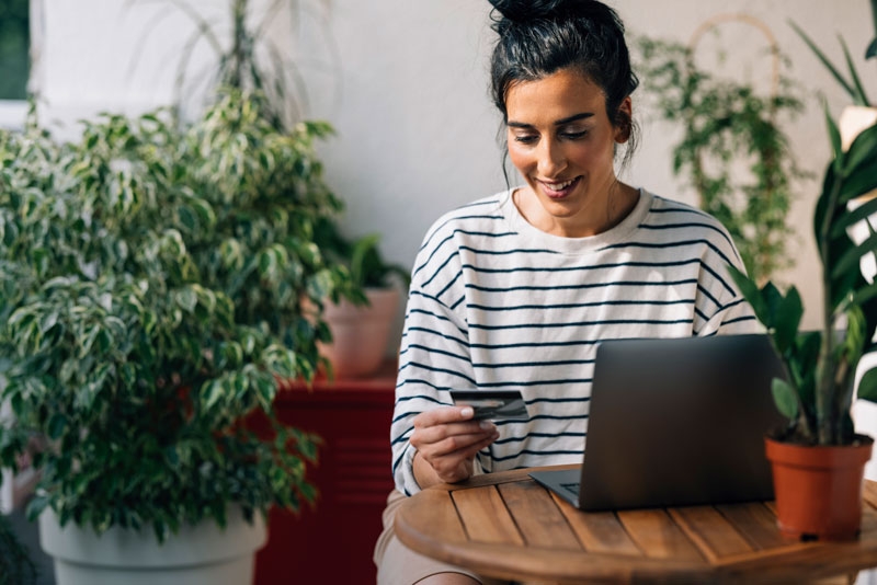 A woman sits at a laptop entering details from a payment card. Behind her are planters with some small plants.