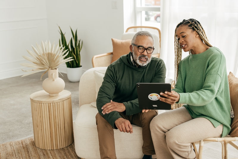 A woman sits with an elderly man on the couch showing him something on an iPad and explaining it to him