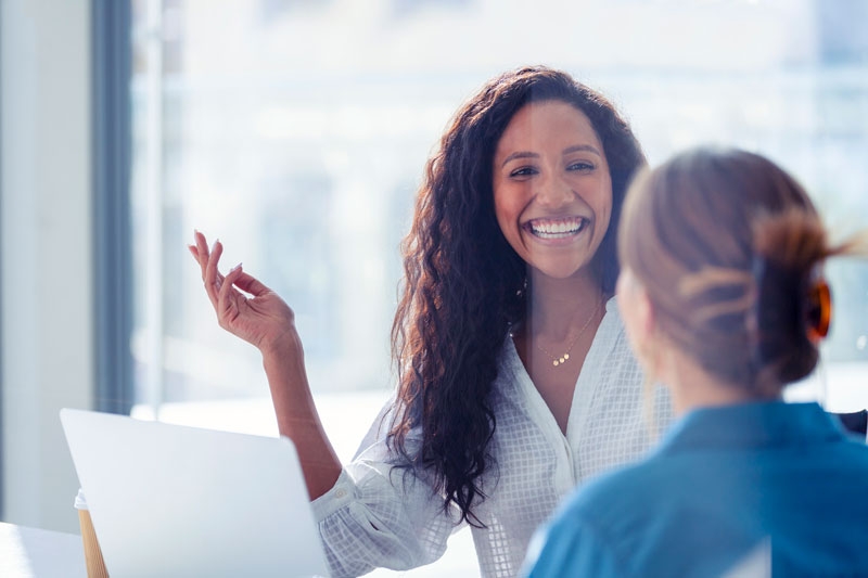 Two women sit in a room together while one explains something from a laptop to the other
