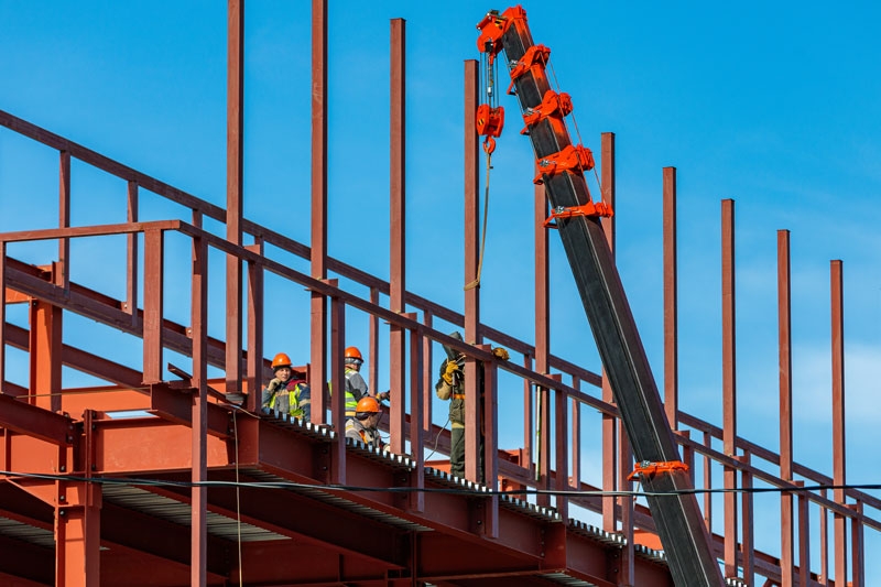 workers are standing on scaffolding as a crane moves beams into position, in the foreground there is a hydro wire